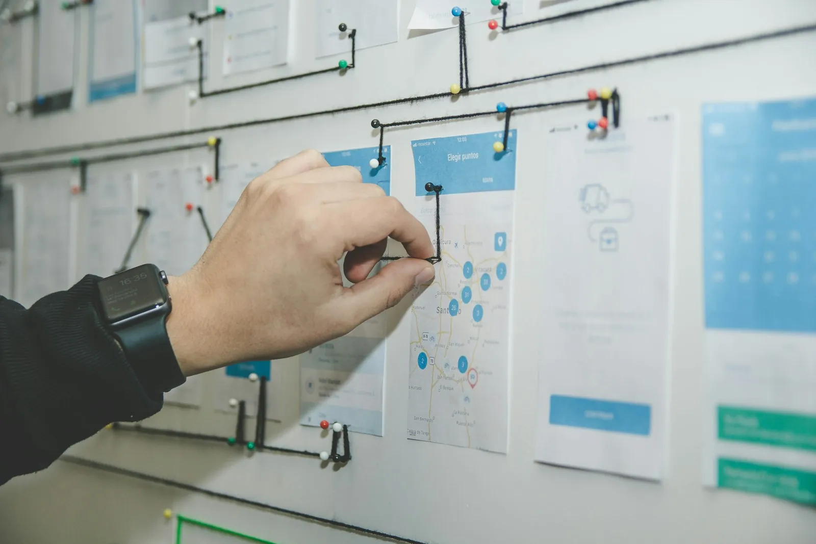 A hand pins printed app and website screen mock-ups to a planning wall linked with thread – planning a website build timeline